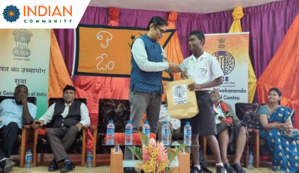 A person on stage is presenting an award or gift bag to a student, with banners in the background indicating involvement from the High Commission of India and the Swami Vivekananda Cultural Centre.