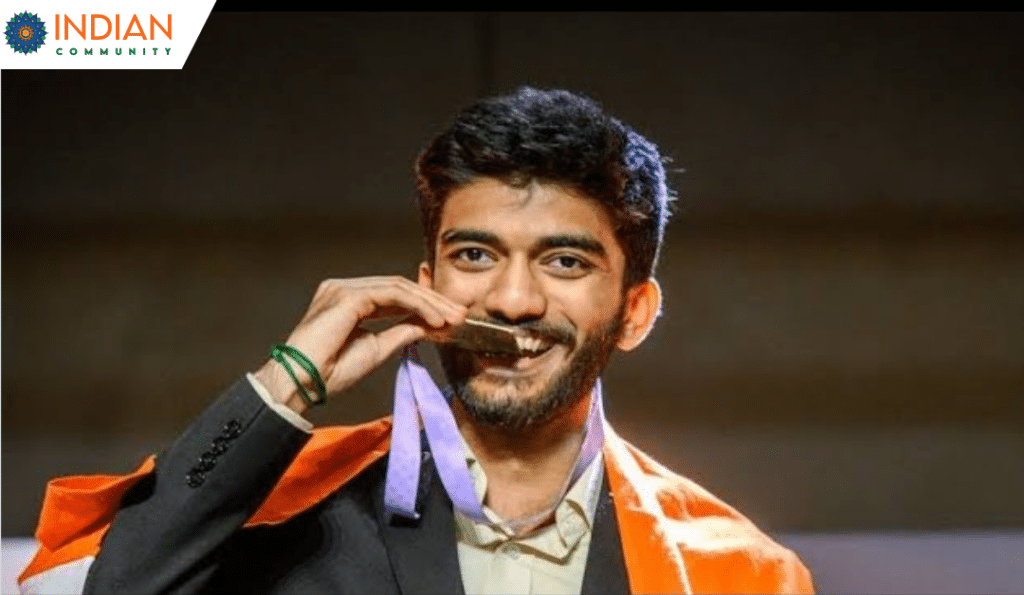 Gukesh Dommaraju in a suit, draped in an Indian flag, bites into a gold medal while smiling.