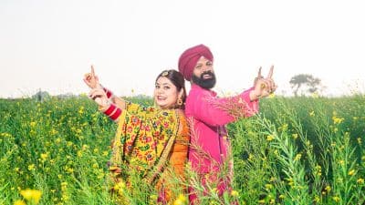Punjabi couple in traditional attire standing in a mustard field, smiling and celebrating with raised hands.