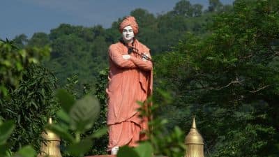 Statue of a Swami Vivekananda in saffron robes standing with folded arms amid greenery and trees.