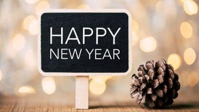 Small sign reading “Happy New Year” placed next to a pine cone against a warm, blurred festive background.